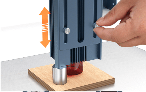 A person's hand adjusting a blue diode laser module to set the correct focal height over a piece of plywood. An orange double-headed arrow indicates vertical movement while a small metal cylinder acts as a focus spacer between the laser housing and the wood surface. This technical demonstration shows how to manually focus a laser for precise cutting and engraving.
