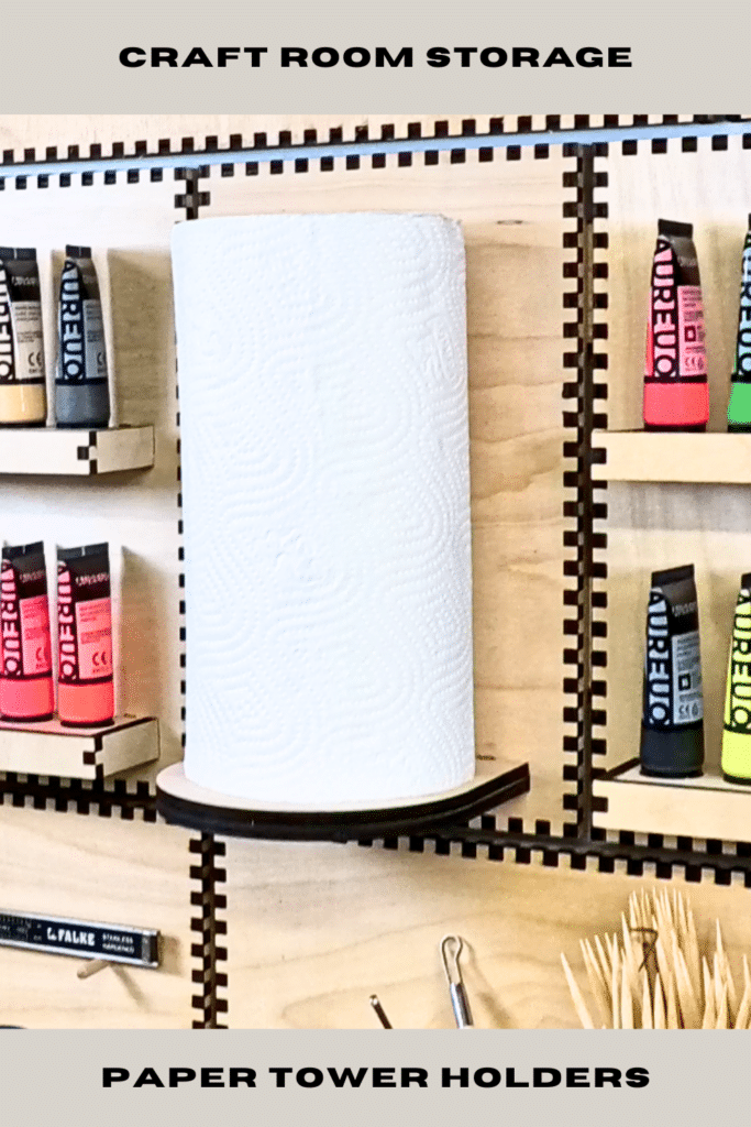 Laser-cut French cleat paper towel holder on a plywood craft wall, holding a vertical white paper towel roll between shelves of colourful acrylic paint tubes and small tools.