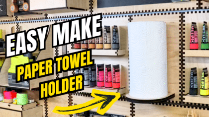 Laser‑cut plywood French cleat paper towel holder mounted on a craft room wall, shown from the front with a white paper towel roll and colourful paint tubes stored on nearby shelves.
