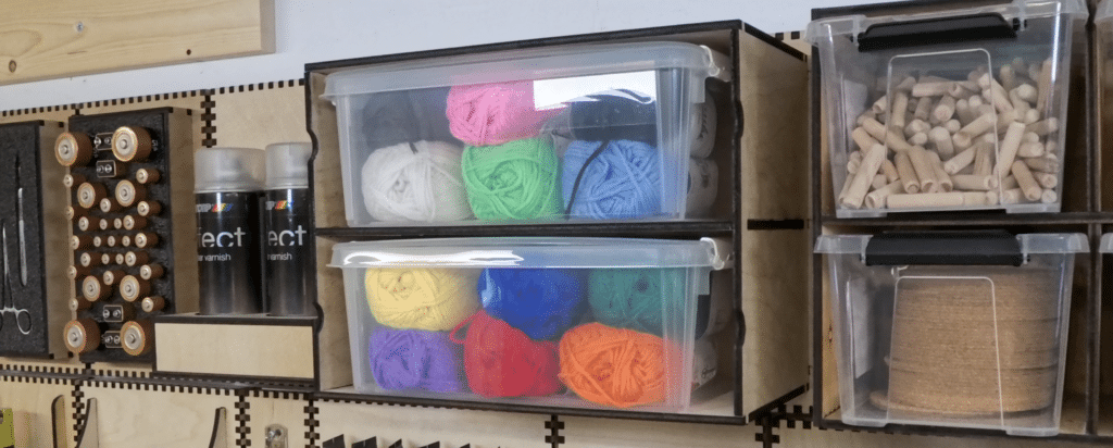 Laser‑cut plywood French cleat cubby holding two clear plastic storage boxes filled with colourful yarn balls, mounted on a workshop wall beside other bin holders.