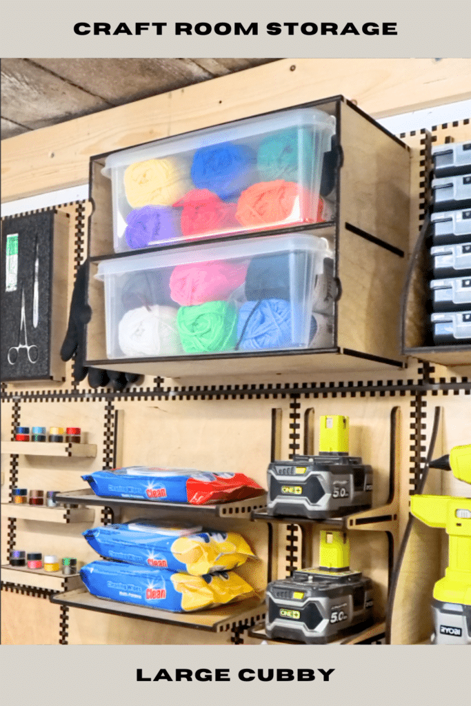 Laser-cut plywood French cleat large cubby holding clear plastic tubs of coloured yarn, cleaning wipes and Ryobi batteries on a modular craft room wall with other small shelves and tools.