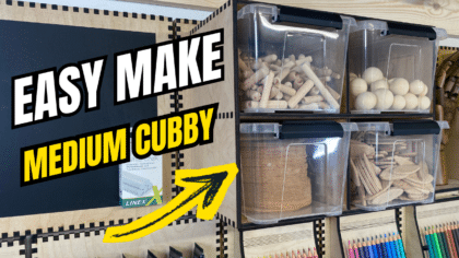 Laser-cut French cleat medium four-box cubby on a plywood wall, holding clear plastic bins filled with wooden dowels, spheres, and biscuits, with bold “Easy Make Medium Cubby” text and a yellow arrow overlay.