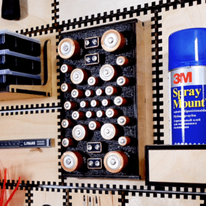 A Neat French Cleat Gallery Frame used for custom battery storage, featuring a black foam insert organized with D, C, AA, AAA, and 9V batteries. The frame is mounted on a modular workshop tool wall with signature interlocking laser-cut plywood joints. A can of 3M Spray Mount is visible to the right, highlighting a professional and efficient workspace organization solution.