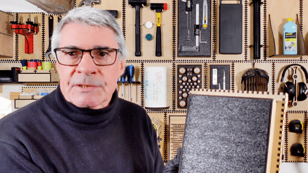 Maker and woodworker Alan presents a new foam-lined, laser-cut component in front of his professional workshop's Neat French Cleat tool wall. The highly organized wall displays a variety of hand tools including mallets, digital calipers, screwdrivers, and safety gear, each secured in custom modular plywood holders. The holders feature characteristic laser-cut interlocking finger joints, highlighting the precision and "Click-Fit" standard of the modular system. This image demonstrates the professional-grade efficiency and aesthetic achievable with precision-engineered workshop organization plans.