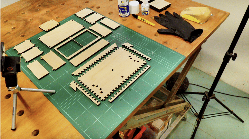 Laser-cut plywood components with intricate interlocking finger joints neatly arranged on a green cutting mat atop a wooden workshop workbench. The organized workspace features assembly essentials like CA glue, "Superfast Activator," a yellow sanding block, and a black protective glove. A camera on a mini-tripod is positioned to film the project, showcasing the precision and "flat-pack" assembly process of the Neat Master System tool holder parts.