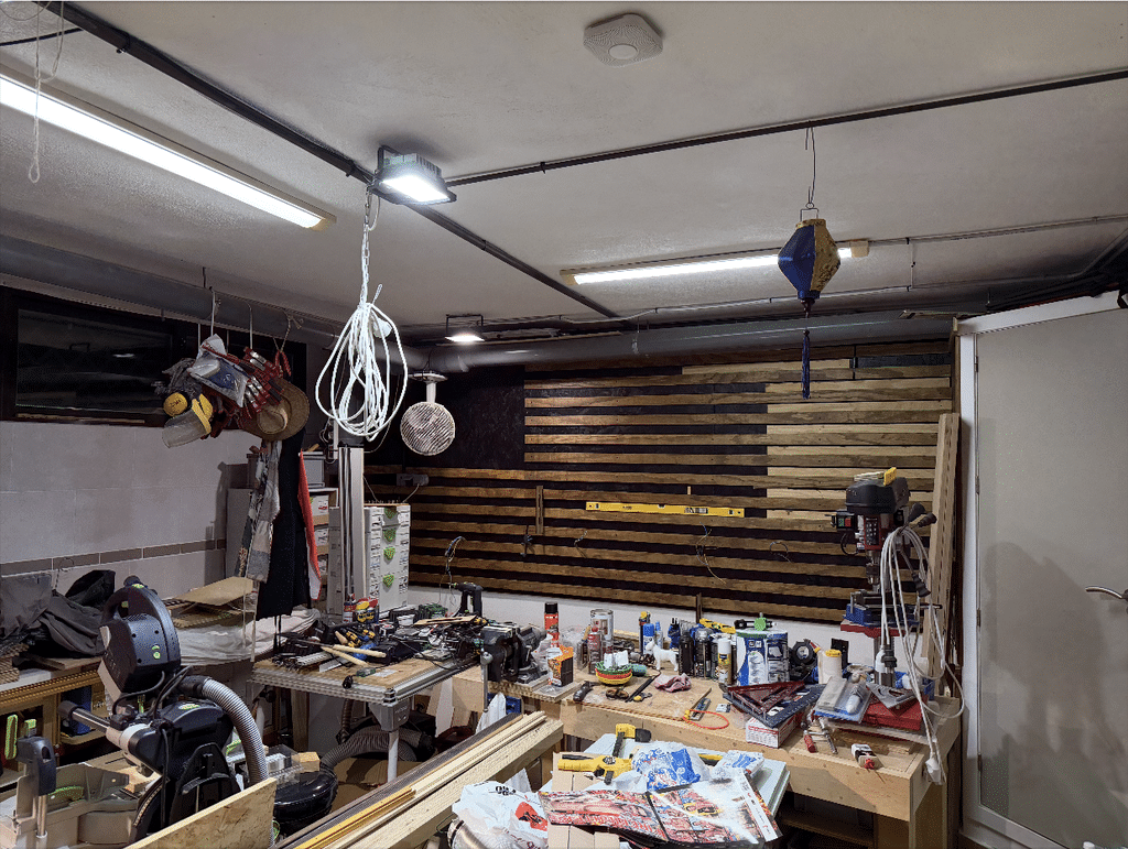 Wide view of Gavin's workshop mid-build, with rows of wall cleats partially installed on the dark-painted back wall above a cluttered workbench of tools, cables, and coiled wiring hanging from the ceiling.