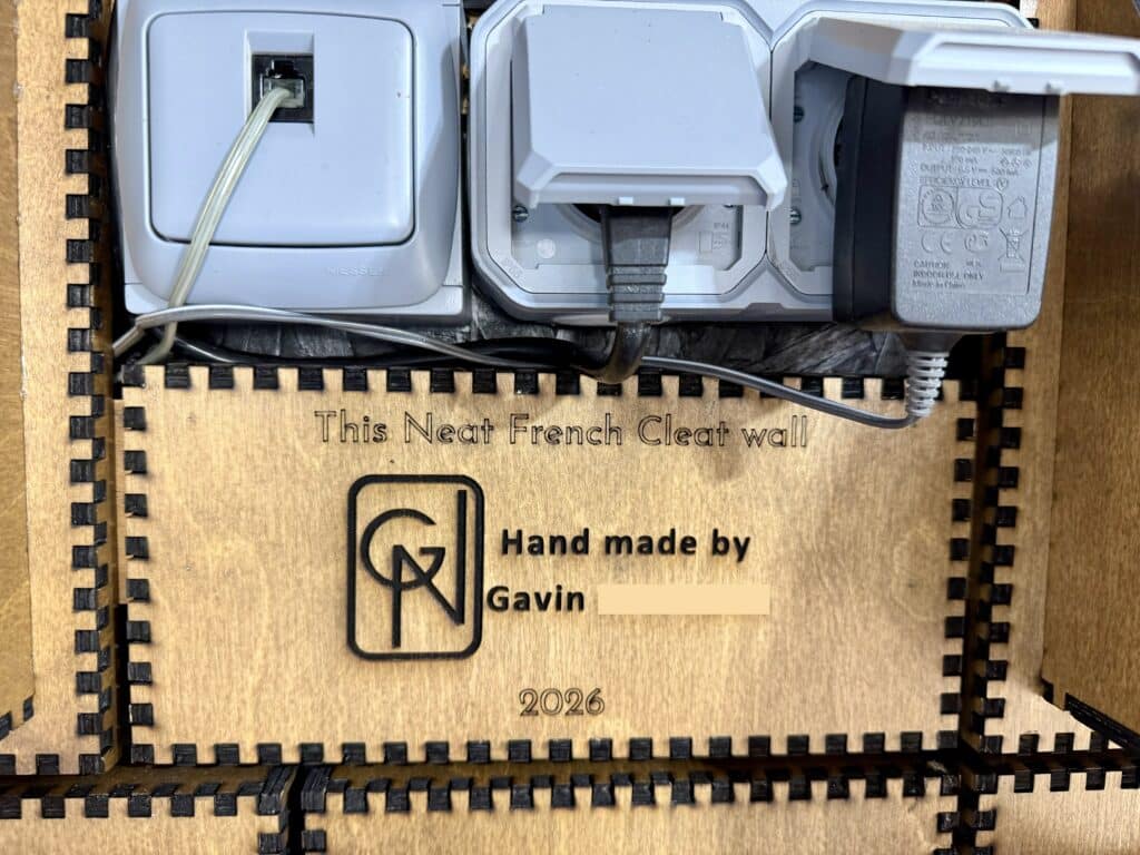 A close-up of the finished cleat wall's built-in electrical and network outlets mounted flush into the panel, with a personalized engraved plaque below reading "This Neat French Cleat wall, Hand made by Gavin, 2026" alongside his logo.