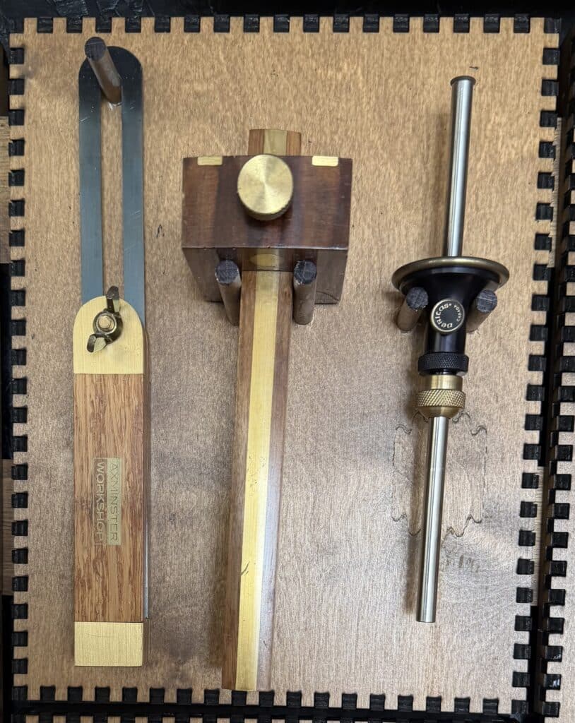 A wooden sliding bevel gauge, a wood-and-brass marking gauge, and a brass wheel marking gauge mounted on a French Cleat panel.