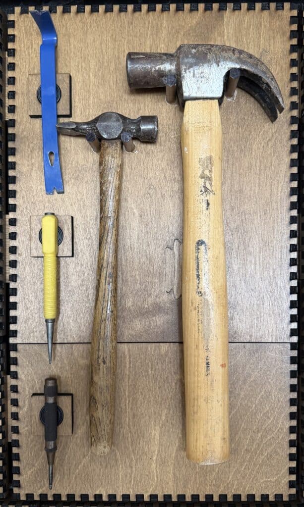 A small tack hammer, a full-size claw hammer, and two awls with blue and yellow handles mounted on a French Cleat panel.