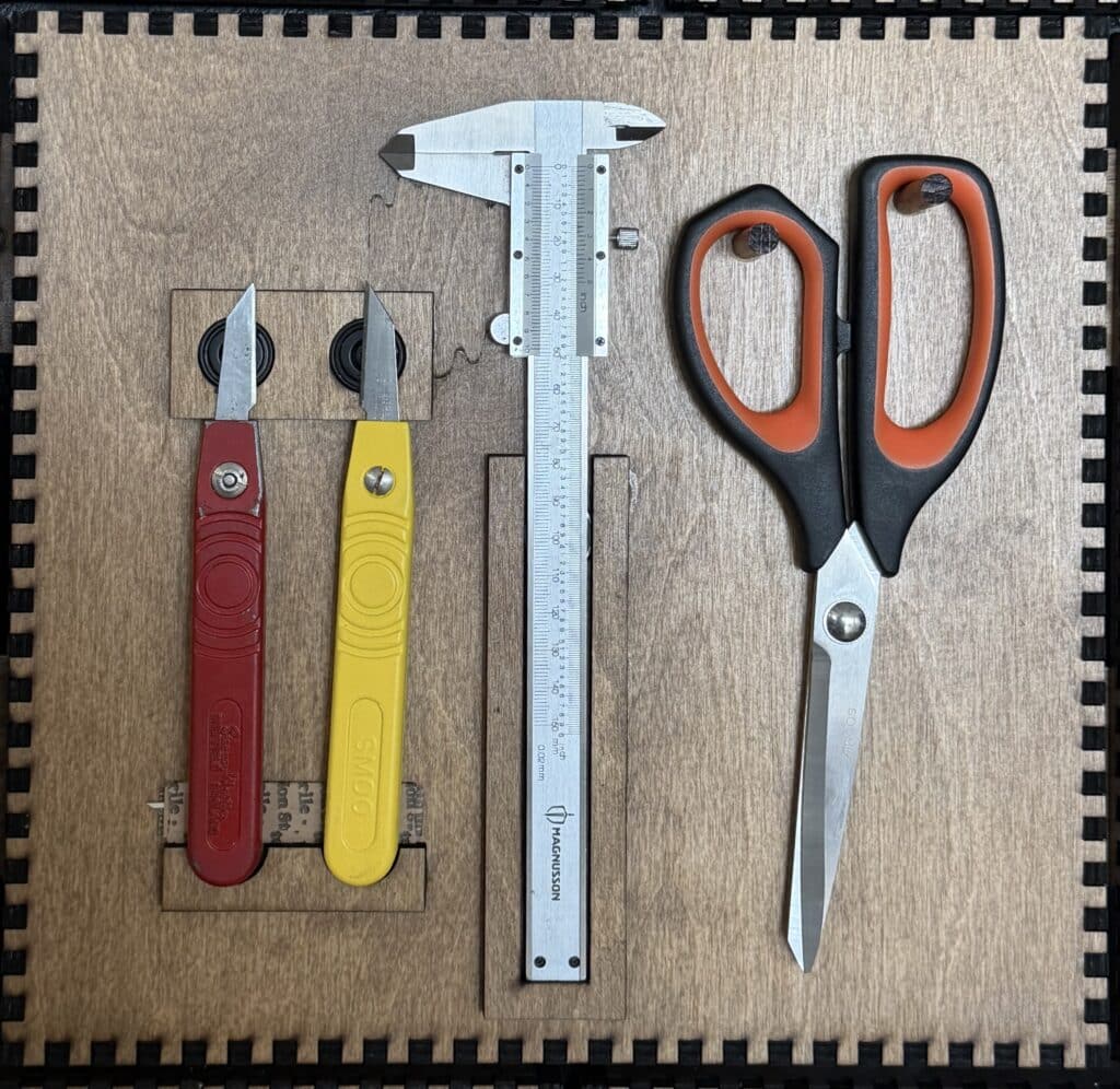 Two folding utility knives, a Magnusson vernier caliper, and a pair of scissors mounted on a French Cleat panel.