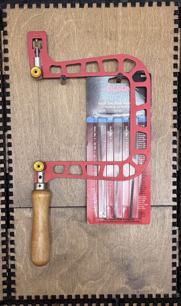 A red-handled coping saw and a mounted pack of OLSON Skip Tooth scroll saw blades hanging on a French Cleat panel.