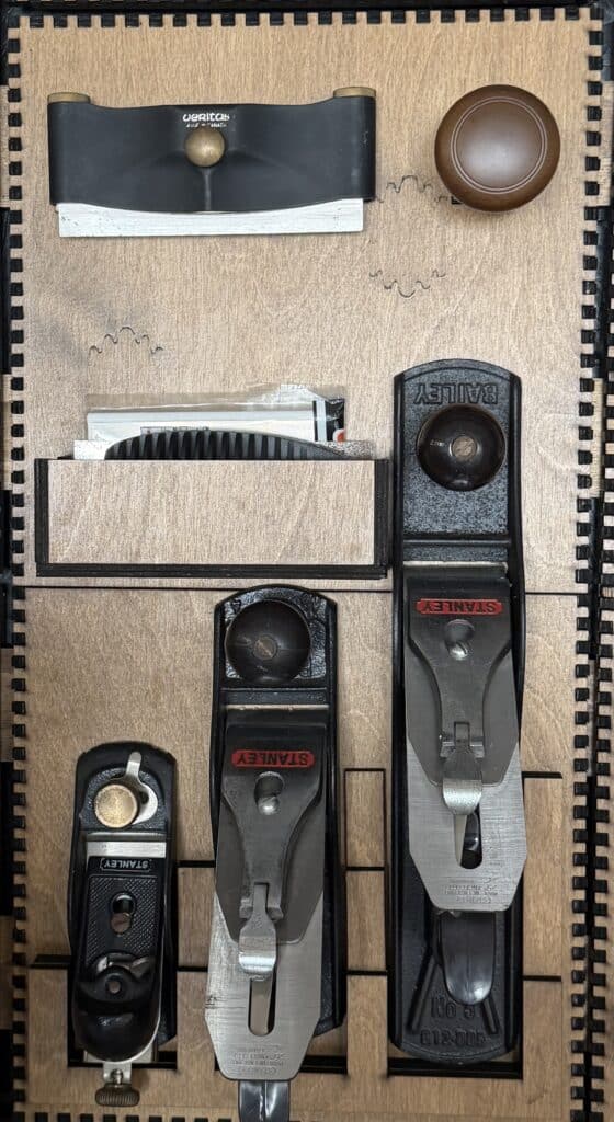 A spokeshave, a wooden knob, stored sandpaper sheets, and three Stanley Bailey hand planes in graduated sizes mounted in a custom laser-cut holder.