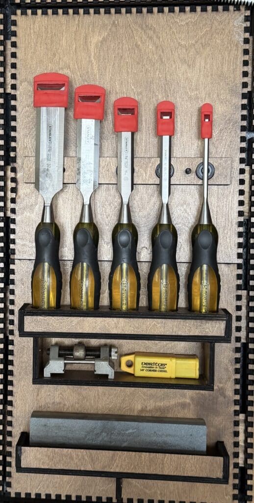 Five graduated wood chisels with red and black handles stored in a custom laser-cut shelf, alongside a honing guide, a small combination square, and a sharpening stone.