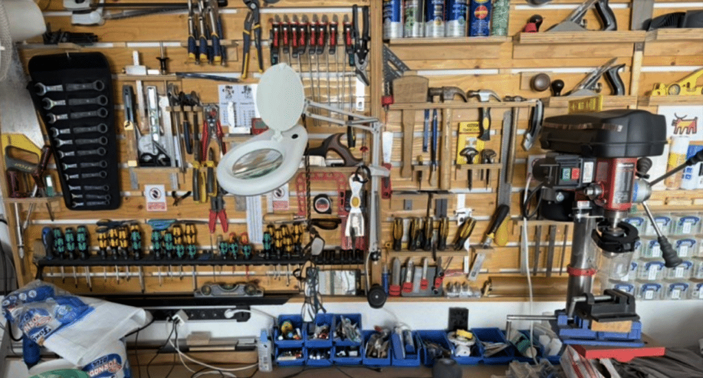 Gavin's original tool wall before the French Cleat system, with wrenches, clamps, screwdrivers, and a magnifying lamp crowded onto basic pegboard hooks, plus a drill press and bins of loose hardware along the floor.