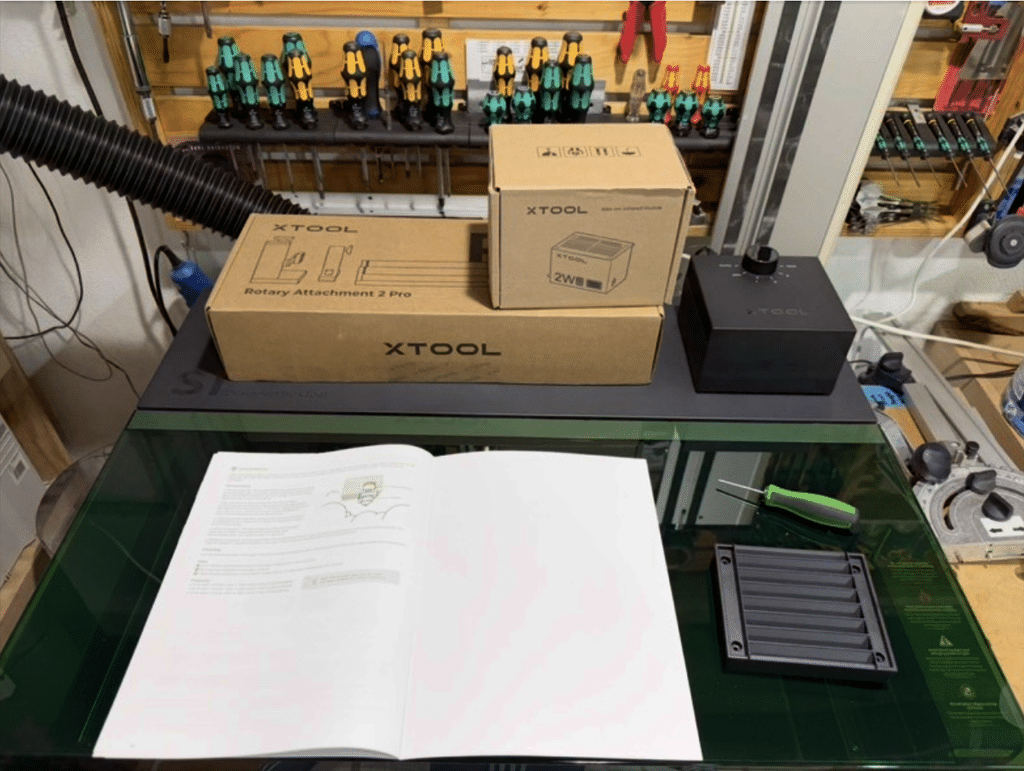 Gavin's xTool laser cutter setup, still boxed on the workbench, with the instruction manual open on the glass bed and a screwdriver set mounted on the pegboard behind it, ready for his first Neat French Cleat cut.