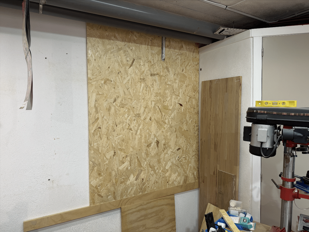 A blank OSB panel mounted on Gavin's basement wall, ready to become his French Cleat system, with a drill press and workbench visible in the corner beside it.