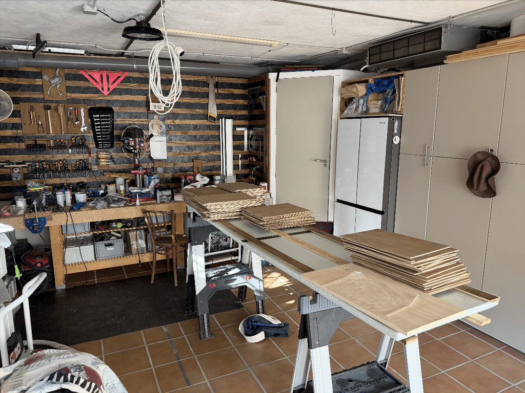 Stacks of freshly cut cleat panels resting on sawhorses in Gavin's workshop, with the partly finished cleat wall and hanging cables visible behind, ready for assembly.