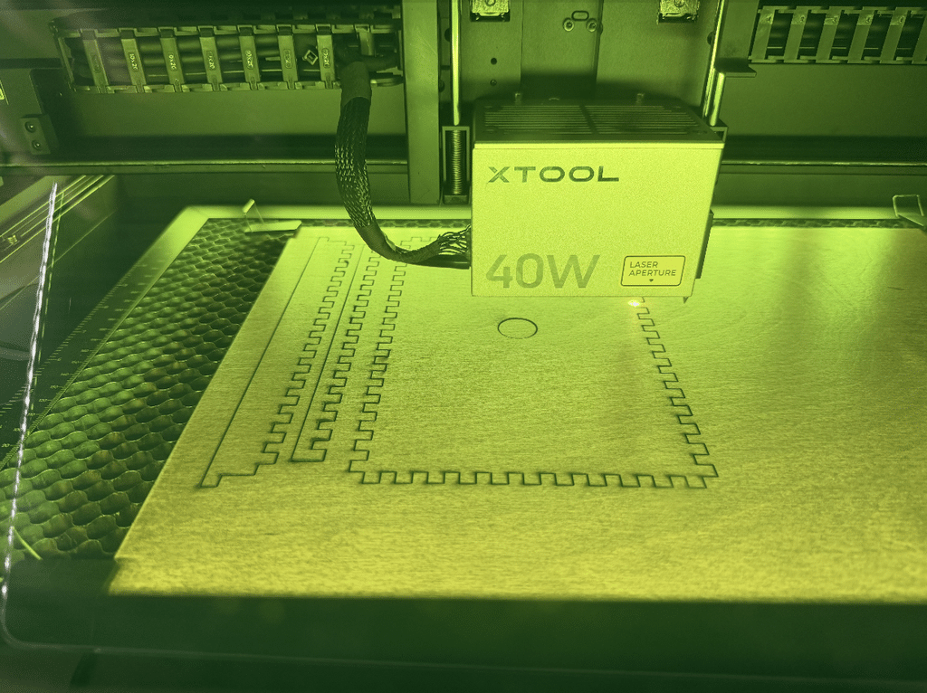 Gavin's xTool 40W laser mid-cut, its green safety light illuminating the machine as it traces the interlocking French Cleat panel pattern into the material.